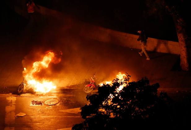 A motorcycle taxi driver walks past burning motorcycles set ablaze by anti-government demonstrators at Altamira Square in Caracas