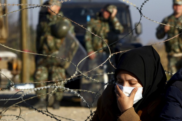 Un migrante cubre su rostro con una máscara médica mientras espera junto a la frontera de Grecia con Macedonia, cerca de la localidad griega de Idomeni, 19 de noviembre de 2015. Los países de los Balcanes han comenzado filtrar el flujo de inmigrantes a Europa, la concesión de pasaje a los que huyen de conflictos en Oriente Medio y Afganistán, pero que dan vuelta la espalda a otros procedentes de África y Asia, las Naciones Unidas y los testigos de Reuters dijeron el jueves. Policías macedonios están en el lado macedonio de la frontera como se ve en el fondo. REUTERS / Alexandros Avramidis