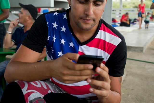 Esta fotografía del viernes 20 de noviembre de 2015 muestra a un inmigrante cubano con una camiseta con un diseño de la bandera de Estados Unidos mientras usa su celular en un refugio en La Cruz, Costa Rica, en la frontera con Nicaragua. Casi todos los cubanos entrevistados en refugios a lo largo de la frontera de Costa Rica con Nicaragua señalaron que los teléfonos inteligentes baratos, ciertos planes de datos y Facebook son el secreto de su éxito para llegar a la frontera con Estados Unidos. (Foto AP/Esteban Félix)