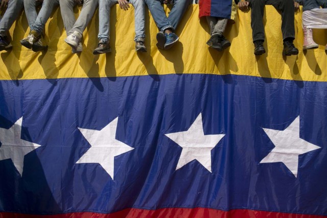 En esta imagen del 26 de octubre de 2016, gente sentada en un muro cubierto por una bandera venezolana durante una protesta contra el presidente Nicolás Maduro en Caracas, Venezuela. La tensión se agravó en Venezuela después de que la Asamblea Nacional votara a favor de abrir un juicio político contra Maduro por romper el orden constitucional, y de que los líderes opositores convocaran marchas masivas el miércoles para expulsar al mandatario.  (AP Foto/Alejandro Cegarra)