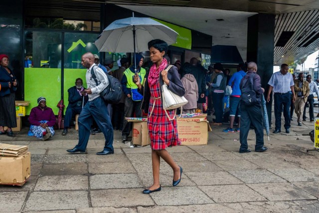 A woman with an umbrella walks past residents queuing outside a bank in Harare on November 15, 2017. Zimbabwe's military appeared to be in control of the country on November 15 as generals denied staging a coup but used state television to vow to target "criminals" close to President Mugabe. / AFP PHOTO / -
