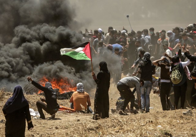 Una mujer palestina sosteniendo su bandera nacional observa los enfrentamientos con fuerzas israelíes cerca de la frontera entre Gaza e Israel al este de la ciudad de Gaza el 14 de mayo de 2018, mientras los palestinos protestan por la inauguración de la embajada de Estados Unidos tras su controvertido traslado a Jerusalén. Docenas de palestinos fueron asesinados por disparos israelíes el 14 de mayo cuando decenas de miles protestaron y estallaron enfrentamientos a lo largo de la frontera de Gaza contra la transferencia de su embajada a Jerusalén, luego de meses de protestas globales, ira palestina y elogios exuberantes de los israelíes por el presidente Donald Trump decisión dejando de lado décadas de precedentes. / AFP PHOTO / MAHMUD HAMS